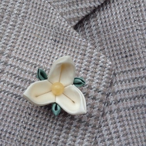 May include: A white fabric flower with a yellow center and green leaves. The flower is pinned to a brown fabric background.