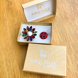 May include: Two lapel pins in open cardboard boxes. One pin is a colorful flower with a pearl center. The other is a small, red and black patterned button. The boxes have the text "EXQUISITE LAPEL" printed on them.