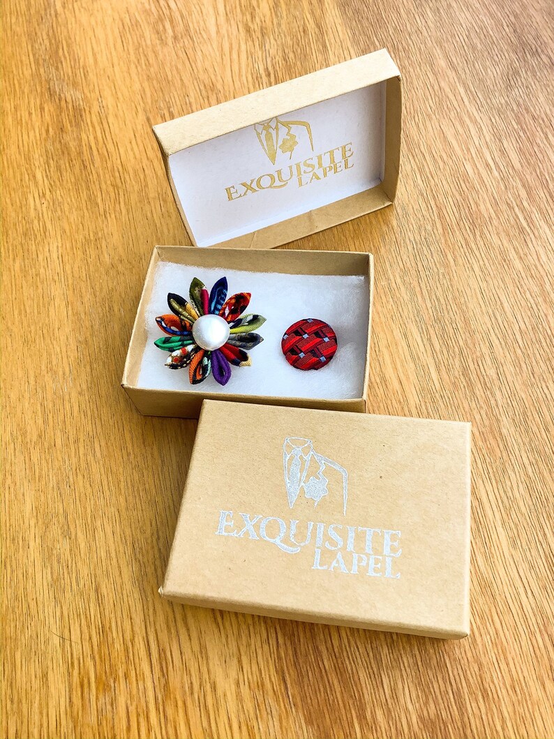 May include: Two lapel pins in open cardboard boxes. One pin is a colorful flower with a pearl center. The other is a small, red and black patterned button. The boxes have the text "EXQUISITE LAPEL" printed on them.