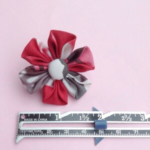 May include: A handmade fabric flower with red, gray, and white patterned petals and a button center. The flower is approximately 2 inches in diameter. A metal ruler with centimeter and inch measurements is next to the flower.