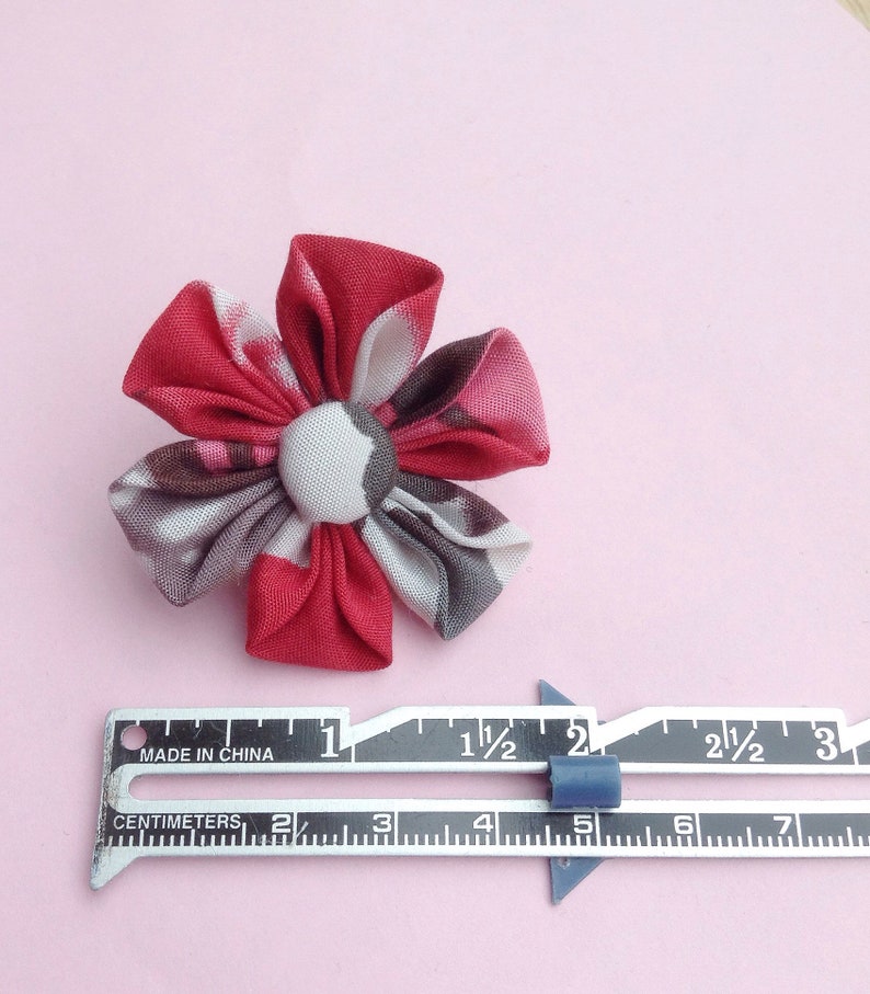 May include: A handmade fabric flower with red, gray, and white patterned petals and a button center. The flower is approximately 2 inches in diameter. A metal ruler with centimeter and inch measurements is next to the flower.