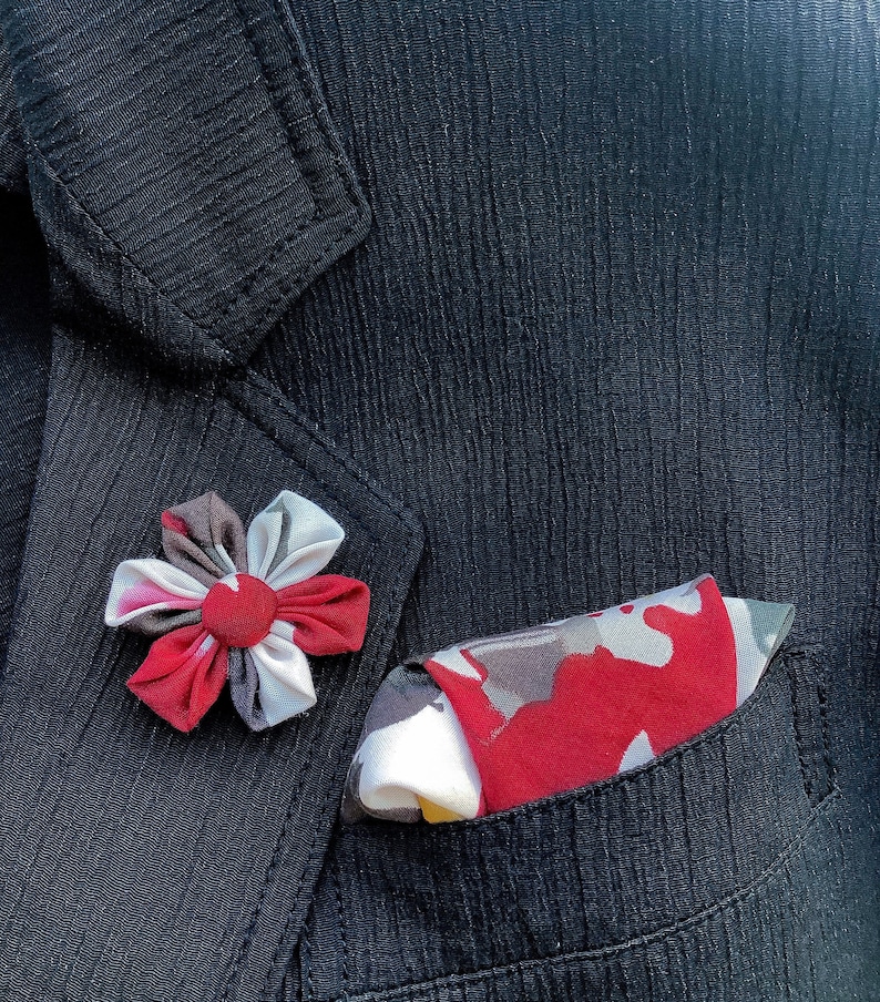 May include: A black blazer with a flower-shaped lapel pin and a pocket square. The pin is red, white, and gray. The pocket square has a red, gray, and white pattern. The blazer has a textured surface.