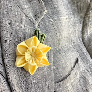 May include: A yellow fabric flower with green leaves, resembling a daffodil, is pinned to a gray fabric background.