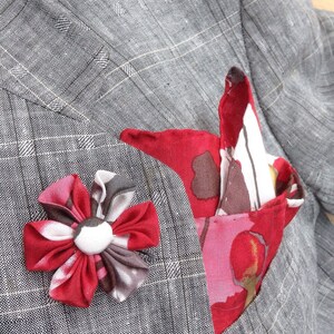 May include: A gray plaid blazer with a red and white floral pocket square and a matching red flower lapel pin. The blazer is made of a textured fabric.