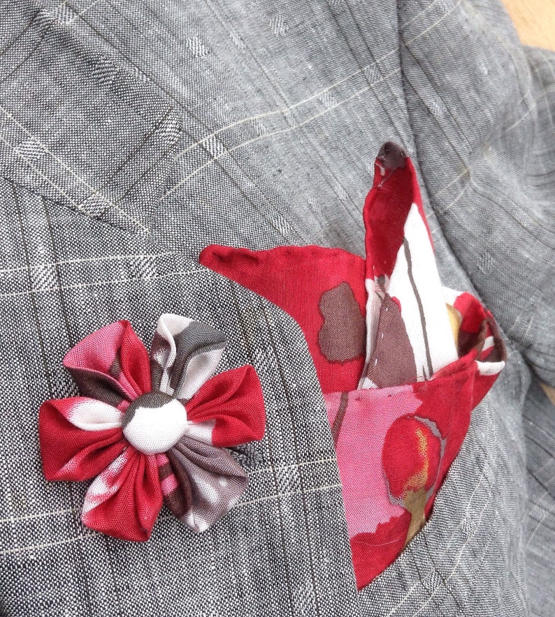 May include: A gray plaid blazer with a red and white floral pocket square and a matching red flower lapel pin. The blazer is made of a textured fabric.