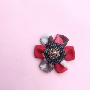 May include: A fabric flower pin with a gold-colored pin back. The flower petals are red, gray, and pink, with a black and gray center. The pin is set against a light pink background.