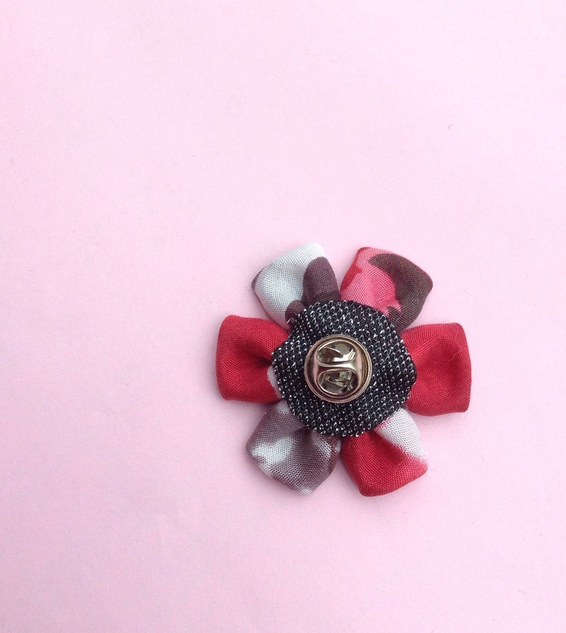 May include: A fabric flower pin with a gold-colored pin back. The flower petals are red, gray, and pink, with a black and gray center. The pin is set against a light pink background.