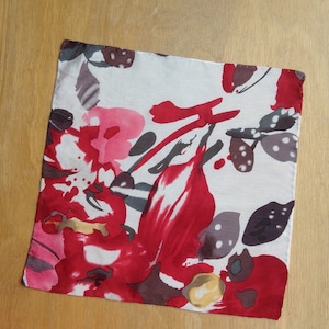 May include: Square white fabric with a vibrant floral pattern in shades of red, pink, gray, and yellow. The design features abstract shapes resembling flowers and leaves. The fabric is likely a handkerchief or a small decorative cloth.