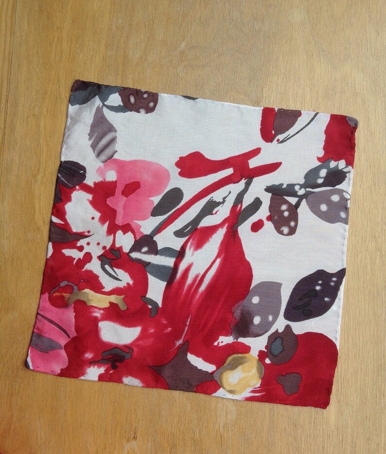 May include: Square white fabric with a vibrant floral pattern in shades of red, pink, gray, and yellow. The design features abstract shapes resembling flowers and leaves. The fabric is likely a handkerchief or a small decorative cloth.