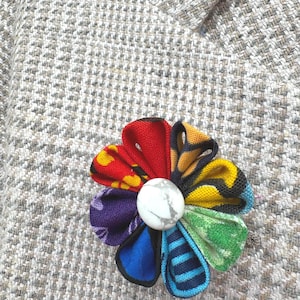May include: A fabric flower brooch featuring eight petals in various colors and patterns, including red, yellow, blue, green, and purple, arranged in a circular shape around a central white stone with grey veining, pinned to a textured beige blazer.