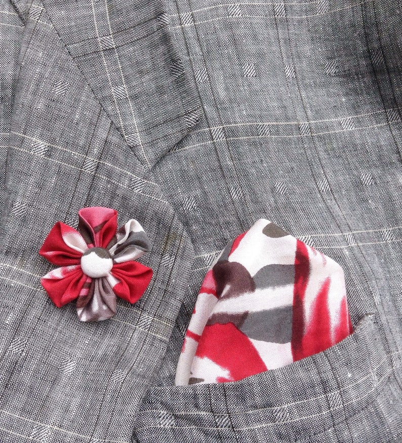 May include: A gray plaid blazer with a red, white, and gray patterned pocket square and a red and gray flower lapel pin. The blazer has a classic design and is made of a textured fabric.
