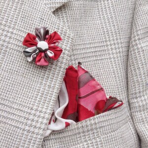 May include: A light gray blazer with a plaid pattern, featuring a red and white floral lapel pin and a red, pink, and white patterned pocket square. The pocket square is folded in a triangle shape.