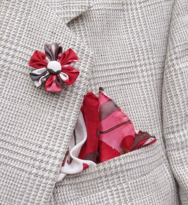 May include: A light gray blazer with a plaid pattern, featuring a red and white floral lapel pin and a red, pink, and white patterned pocket square. The pocket square is folded in a triangle shape.