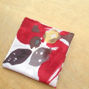 May include: Folded square scarf with a floral pattern. The scarf features a white background with large red, brown, and yellow abstract floral designs. The scarf is folded neatly on a light wood surface.