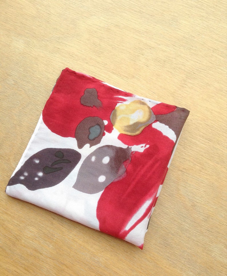 May include: Folded square scarf with a floral pattern. The scarf features a white background with large red, brown, and yellow abstract floral designs. The scarf is folded neatly on a light wood surface.