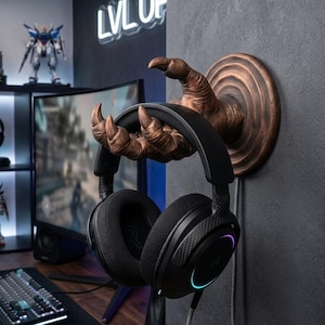 May include: A bronze claw-shaped headphone holder mounted on a dark grey wall, holding black over-ear headphones. The headphones have a multi-coloured light accent. The background shows a gaming setup with a keyboard and monitor.