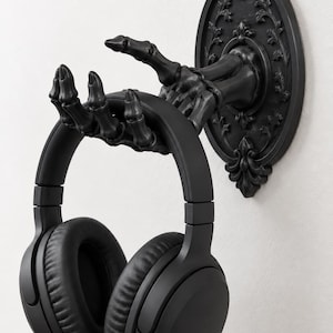 May include: A matte black wall-mounted headphone stand shaped like a skeletal hand extending from an oval, ornate, floral-patterned backplate. The fingers of the skeletal hand are curved upward to securely hold a pair of solid black over-ear headphones.