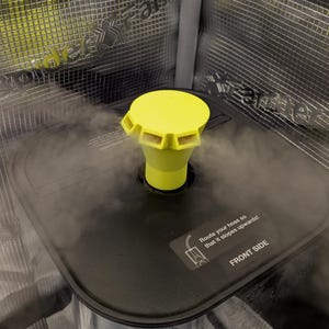 May include: A close-up of a black rectangular object with a yellow nozzle and a yellow cap. The object is emitting steam. The text "Route your hose so that it slopes upwards! FRONT SIDE" is visible on the object.