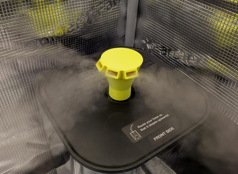 May include: A close-up of a black rectangular object with a yellow nozzle and a yellow cap. The object is emitting steam. The text "Route your hose so that it slopes upwards! FRONT SIDE" is visible on the object.