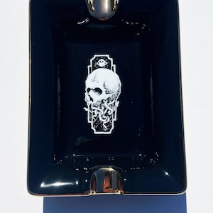 May include: Black ceramic ashtray with a gold rim and a white graphic of a skull with an eye and tentacles.