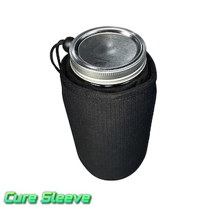 Puede incluir: Funda aislante negra con tapa plateada. La funda está diseñada para contener un tarro. El texto "Cure Sleeve" se muestra en verde en la parte inferior de la imagen. La funda tiene un cierre de cordón.