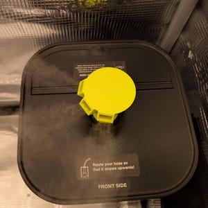May include: A black, square-shaped device with a bright yellow cap. The device has a sticker that reads "Route your hose so that it slopes upwards! FRONT SIDE". Steam is visible, suggesting it is in operation.