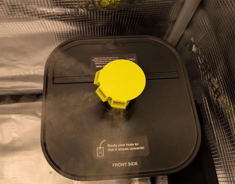 May include: A black, square-shaped device with a bright yellow cap. The device has a sticker that reads "Route your hose so that it slopes upwards! FRONT SIDE". Steam is visible, suggesting it is in operation.