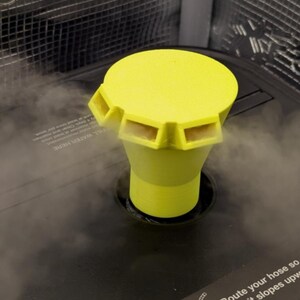 May include: A bright yellow humidifier cap with a cylindrical base and a flat top with four rectangular vents. The cap is set into a dark gray humidifier base, with steam visible around the cap. The text "FILL WATER HERE" is partially visible.