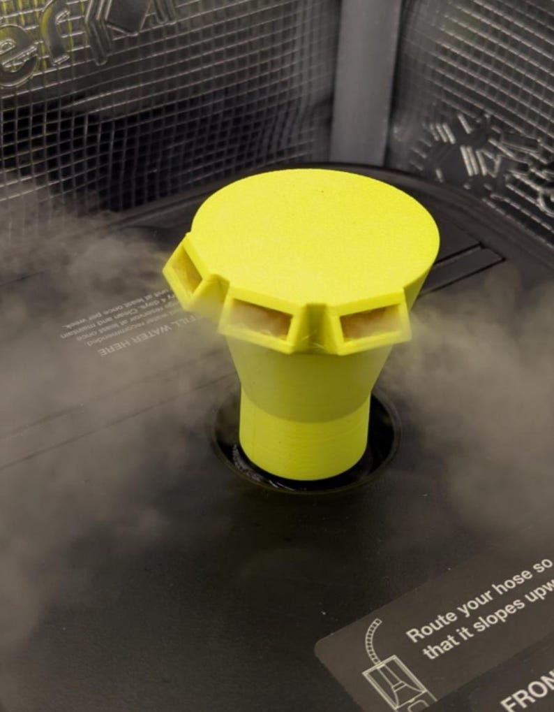 May include: A bright yellow humidifier cap with a cylindrical base and a flat top with four rectangular vents. The cap is set into a dark gray humidifier base, with steam visible around the cap. The text "FILL WATER HERE" is partially visible.