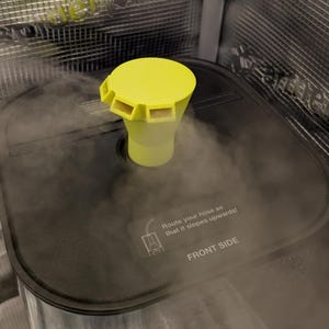 May include: A yellow and clear plastic device with a cylindrical base and a flared top with rectangular openings. The device is positioned on a black surface, with steam rising around it. The text "Route your hose so that it slopes upwards!" is visible.