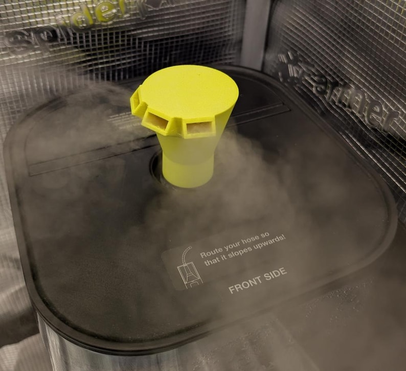 May include: A yellow and clear plastic device with a cylindrical base and a flared top with rectangular openings. The device is positioned on a black surface, with steam rising around it. The text "Route your hose so that it slopes upwards!" is visible.