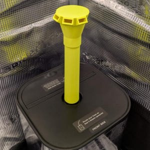 May include: A black square base with a yellow, cylindrical component. The yellow component has a flared top with rectangular openings. The base has a label that reads "FRONT SIDE". The background is a metallic mesh.