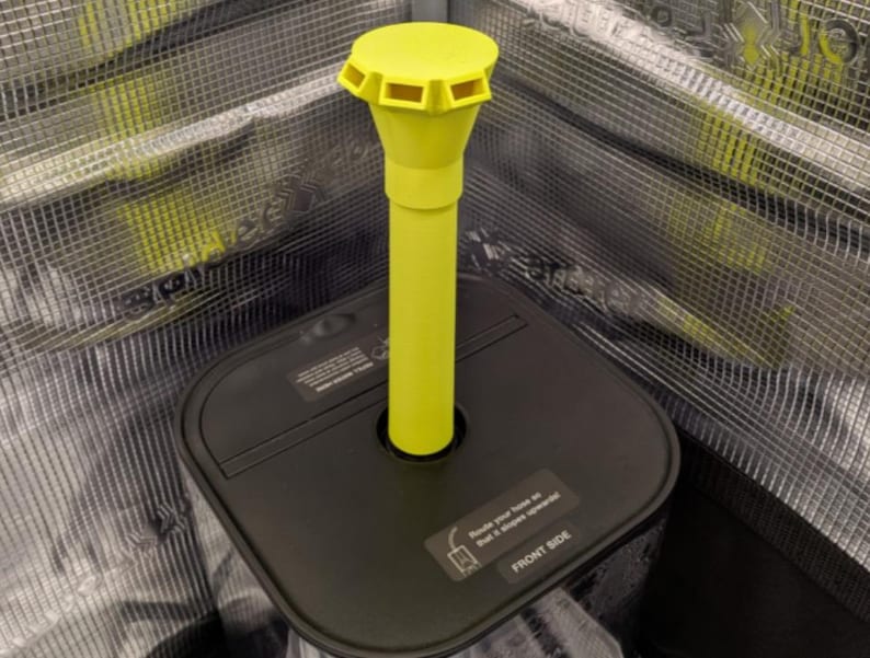 May include: A black square base with a yellow, cylindrical component. The yellow component has a flared top with rectangular openings. The base has a label that reads "FRONT SIDE". The background is a metallic mesh.