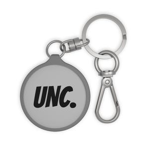 May include: A gray keychain with a round tag featuring the black letters "UNC." The keychain has a silver-colored split ring and a lobster claw clasp for easy attachment to keys or bags.