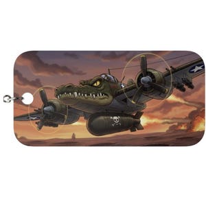 Può includere: Un portachiavi rettangolare con un'illustrazione a cartoni animati di un aereo militare con la testa di un coccodrillo. L'aereo è verde oliva con una bomba con teschio e ossa incrociate, su uno sfondo di tramonto.