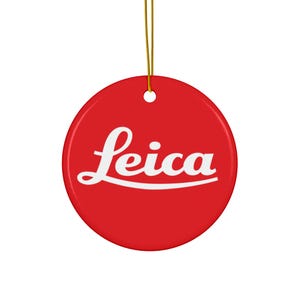 May include: A round, red ornament with the white "Leica" logo. The ornament is suspended by a gold string, ready for hanging. The design is simple and bold, perfect for photography enthusiasts.