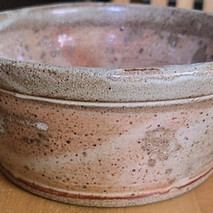May include: A handmade ceramic bowl with a rustic aesthetic. The bowl has a wide, rounded shape with a speckled, earthy colour palette of beige, brown, and pink. The rim has a textured, slightly rough appearance.