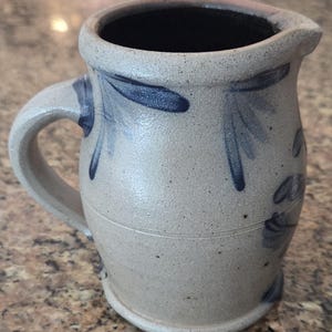 Può includere: Brocca in gres con manico e beccuccio, di colore grigio chiaro e motivi floreali blu. La brocca ha un interno scuro e una superficie strutturata, adatta per servire bevande.