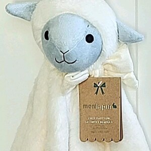 May include: A plush security blanket featuring a soft white fabric body and a light blue lamb head with dark embroidered eyes and a nose. A brown cardboard tag is attached, displaying the brand name monlapin and the text THE LAMB LOP SECURITY BLANKET.