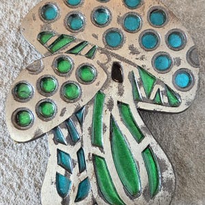 Peut inclure: Broche en forme de champignon argenté avec des accents d'émail vert et turquoise. Le chapeau du champignon présente des cercles turquoise, tandis que le pied et les branchies ont des motifs géométriques verts et turquoise. La broche a un aspect vintage.