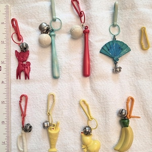 May include: A selection of vintage keyrings with charms such as a deer, baseball bat, fan, and banana. Each keyring has a small bell and a clip. The keyrings are in red, blue, yellow, and white. The ruler shows measurements in inches.