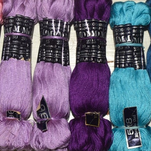 May include: A selection of embroidery threads in shades of pink, lilac, purple, turquoise, and blue. Each skein is labelled with brand details, colour numbers, and length, with some labels showing 25 metres or 27.3 yards.