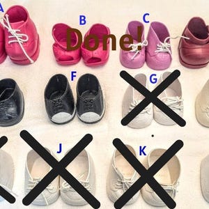 May include: A collection of doll shoes labeled A through L, including bright pink lace-up sneakers, pink open-toe sandals, light pink lace-up shoes, metallic pink shoes, black lace-up shoes, black and white sneakers, and off-white lace-up shoes.