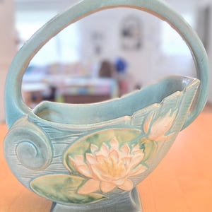 Peut inclure: Un vase en céramique bleu clair en forme de panier, avec une anse. Il présente un motif en relief d'un nénuphar avec une fleur crème et des feuilles vertes. Le vase repose sur une base carrée.