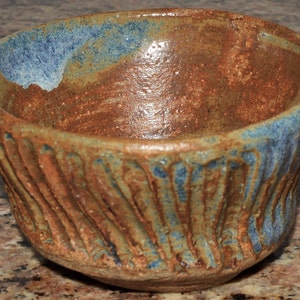 May include: A handcrafted ceramic bowl with a rustic appearance. The bowl has a textured, ridged exterior and a colour palette of brown, blue, and tan. The interior has a smooth finish, while the rim is accented with a blue glaze.