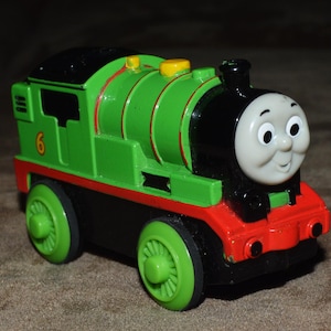 May include: A green toy train with black wheels and a black roof. The train has a smiling face and the number 6 on the side. A children's toy train.