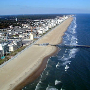 May include: An aerial view of a long sandy beach coastline featuring a wooden pier extending into the dark blue ocean. A paved promenade runs parallel to the shore, lined with numerous multi-storey hotels and buildings under a clear blue sky.