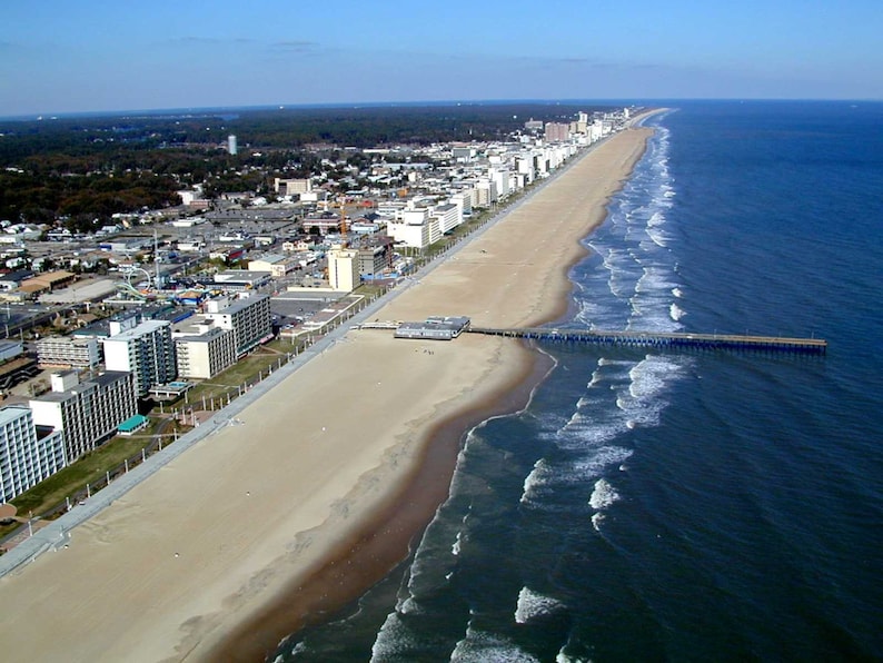 May include: An aerial view of a long sandy beach coastline featuring a wooden pier extending into the dark blue ocean. A paved promenade runs parallel to the shore, lined with numerous multi-storey hotels and buildings under a clear blue sky.