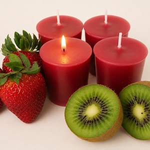 May include: Four cylindrical red votive candles, one of which is lit with a visible flame, arranged alongside two whole red strawberries with green leafy tops and two halves of a green kiwi fruit showing black seeds and a light center.