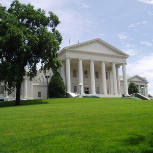 May include: A large white neoclassical building with a prominent triangular pediment supported by tall columns. The structure features multiple rectangular windows and is situated behind a wide, vibrant green lawn with a large tree on the left side.